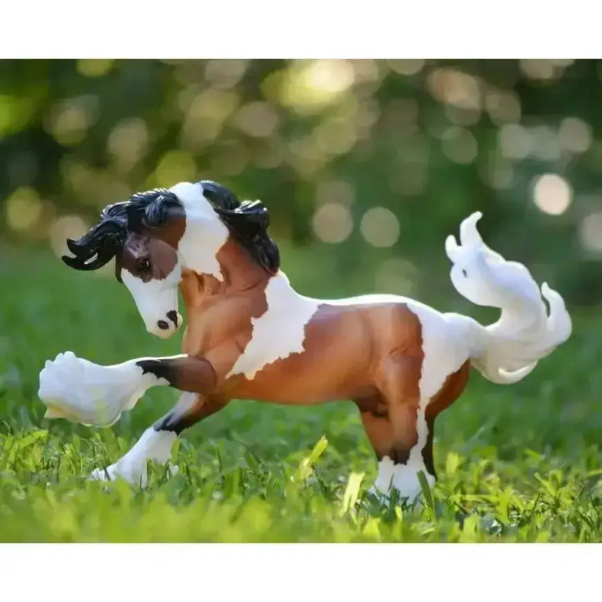 A breyer gypsy vanner model horse with a brown and white pinto coat standing in grass inspired by local traveler communities