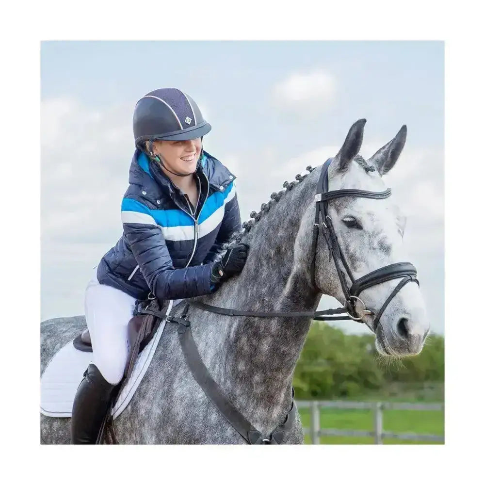 A smiling rider wears a coldstream southdean quilted jacket navy white blue riding a dapple grey horse with black tack
