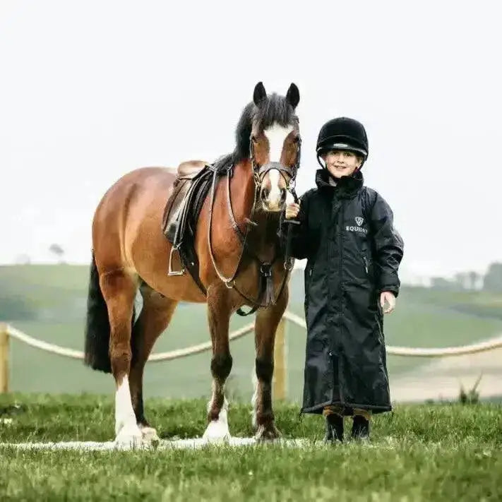 A young child wearing a black equidry equimac waterproof riding jacket with mesh lining and taped seams stands by a horse