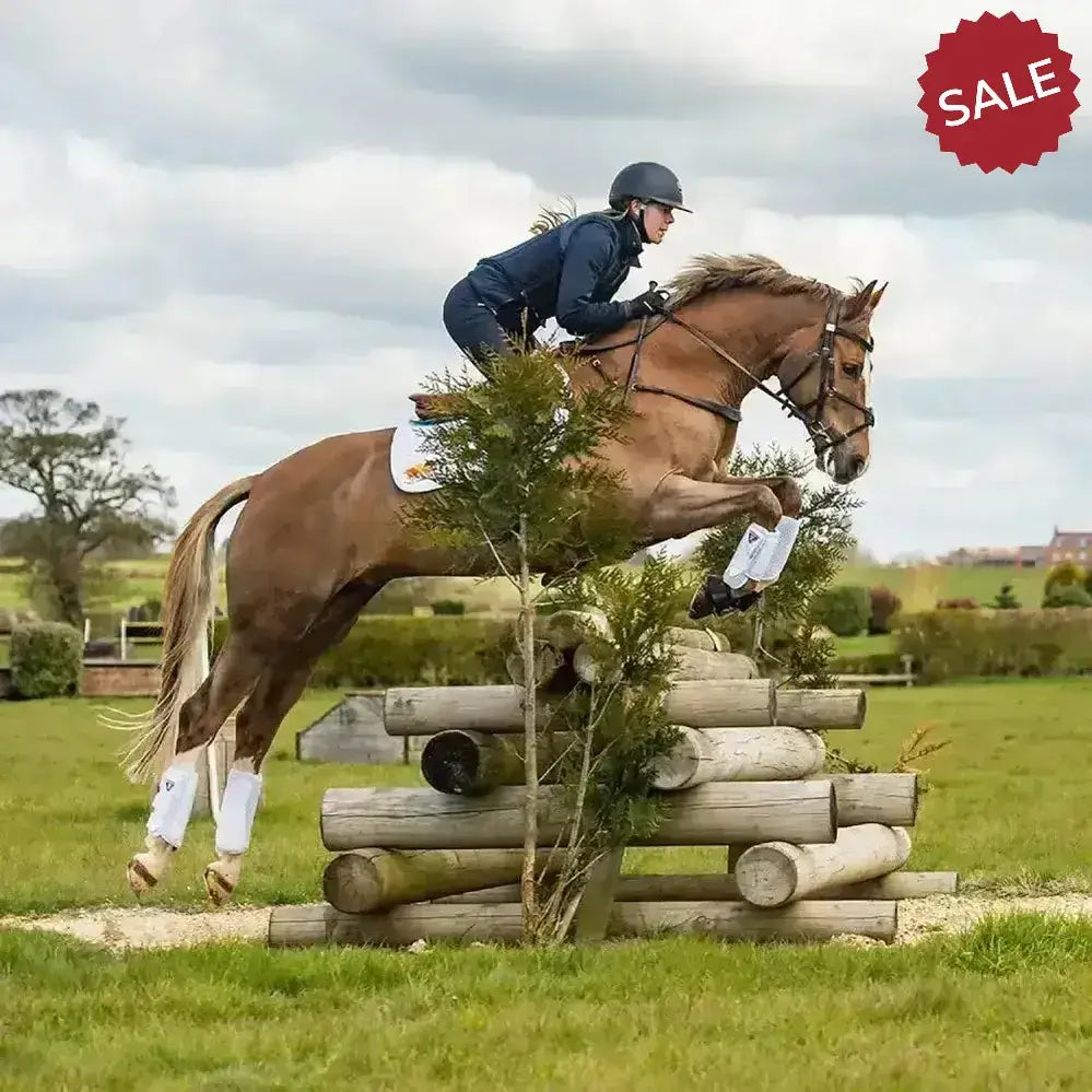 A chestnut horse mid-jump wearing white equilibrium tri-zone cross country impact sports boots on its legs
