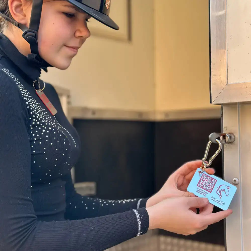 Light blue rectangular equine emergency tag with qr code and horse logo attached to metal carabiner for instantly accessible emergency contact details