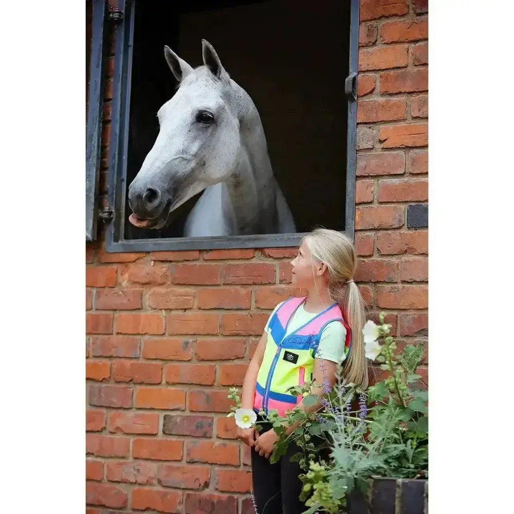 A young girl wearing equisafety childrens hi-vis waistcoats multi-coloured stands beside a brick stable window