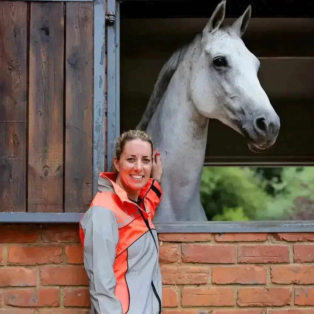 A woman wearing a gray and orange equisafety mercury lightweight reflective riding coat stands by a brick stable wall