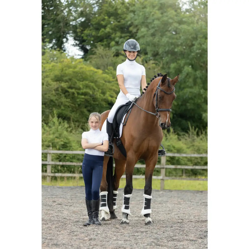A chestnut horse with black and white leg wraps stands next to a hy equestrian children’s roka regal show shirt in an arena