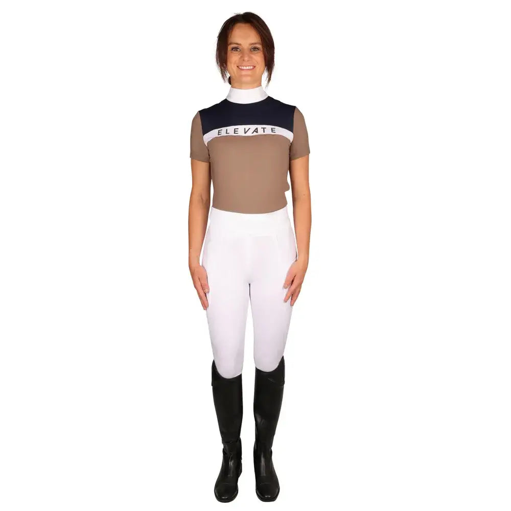 A woman wearing a beige and navy blue hy equestrian elevate allure short sleeve show shirt with white riding pants and black boots