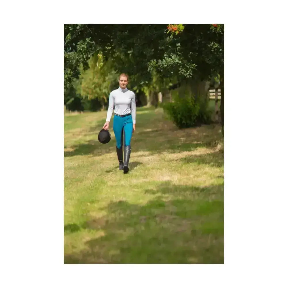 A woman walks holding a black riding helmet wearing hy equestrian hyedition full seat riding breeches on a grassy path