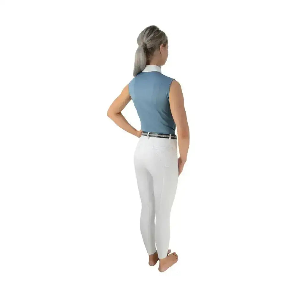 A woman wearing a light blue hy equestrian sophia sleeveless show shirt with silk trim detailing and white riding pants