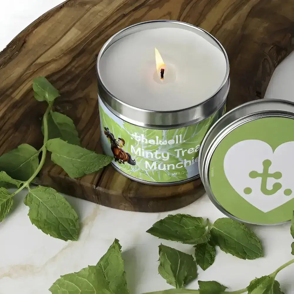 A lit white scented candle in a silver tin with a minty tree munchie label and fresh mint leaves from hy equestrian thelwell collection long-lasting aromatic candle