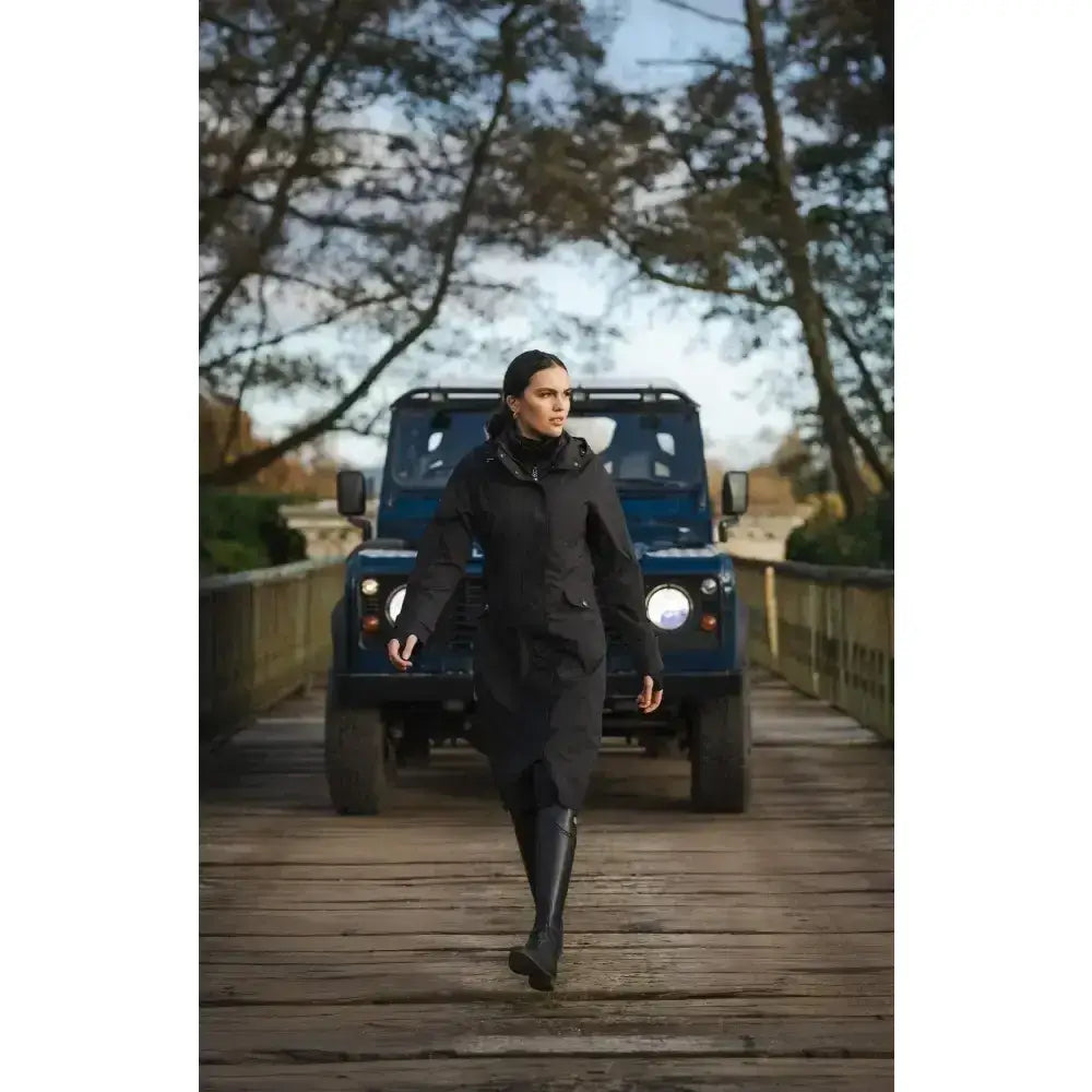 A woman strides across a bridge wearing a black lemieux amelie waterproof lightweight riding coat with tall boots