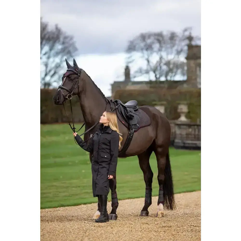 A dark brown horse stands beside a woman wearing the lemieux amelie waterproof lightweight black riding coat