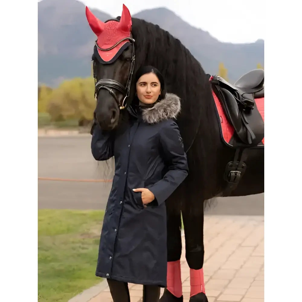 A woman in a navy lemieux anya waterproof faux fur riding coat stands next to a black horse with red gear and pink wraps