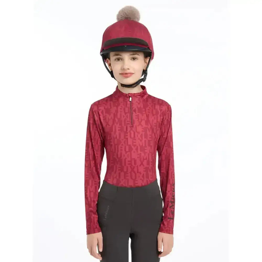 A young girl wears a vibrant red lemieux casey hat silk ember riding helmet with a fluffy pom-pom and black trim