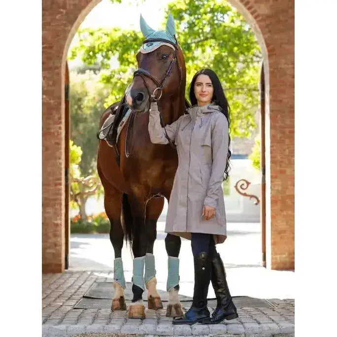 A woman in riding gear stands next to a brown horse wearing blue accessories while wearing a lemieux grace long rain jacket pecan