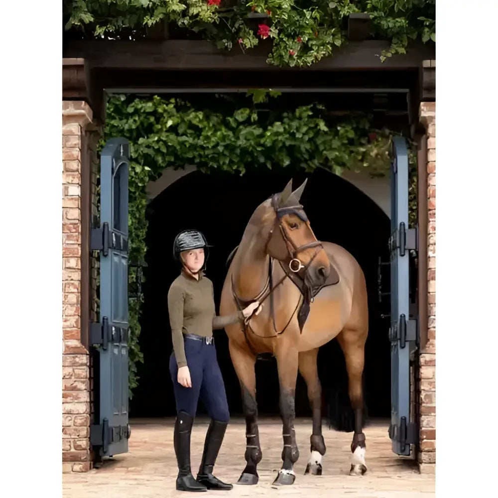 A tan horse with black bridle and leg wraps stands beside a rider wearing the lemieux halle lightweight base layer alpine
