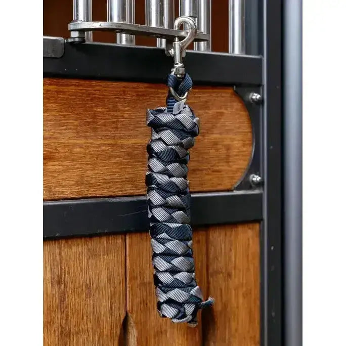 A braided black and gray rope halter strap with metal hook hanging from a stable door lemieux harlequin leadrope navy pecan