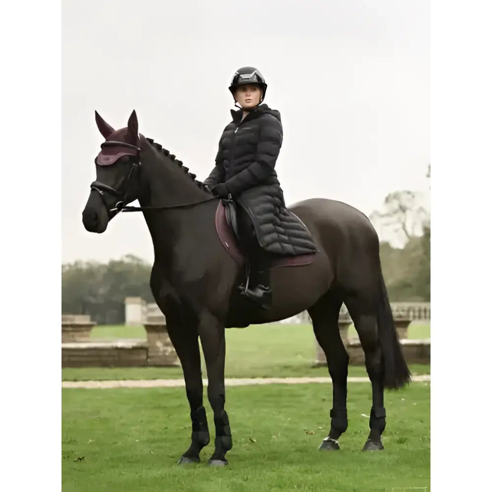 A rider wearing a lemieux hayley water resistant longline puffer coat in black sits on a dark brown horse with matching bridle