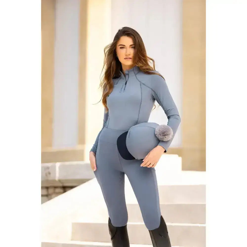 Light blue lemieux ladies base riding suit with zipper neckline and long sleeves paired with black boots layer jay blue