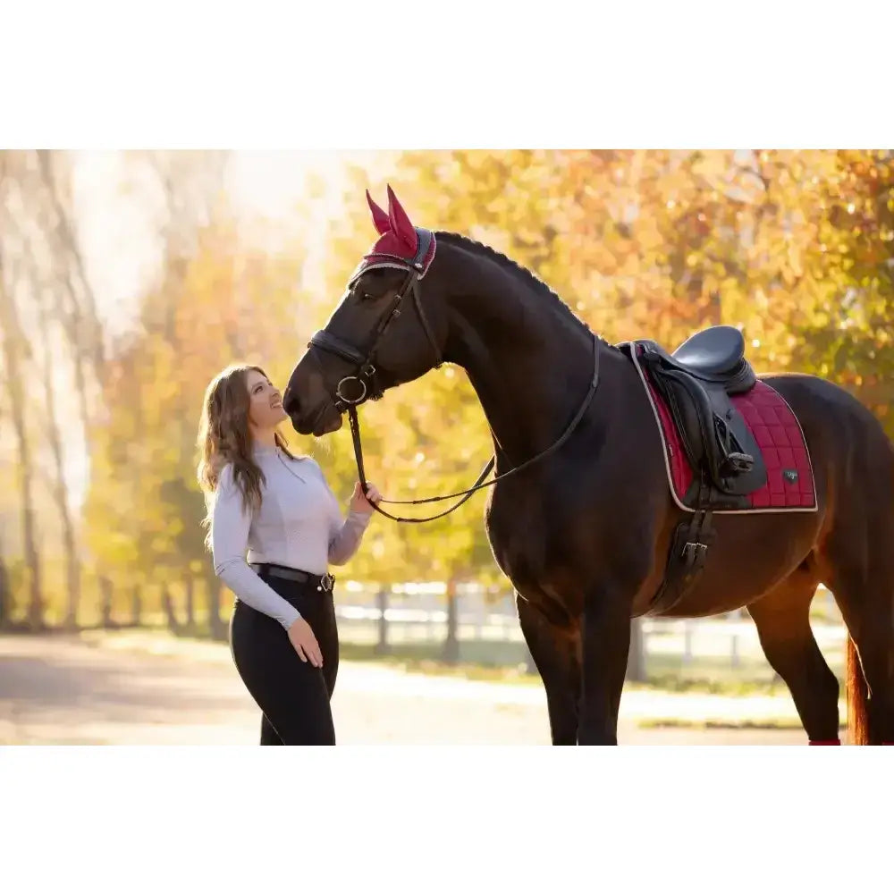 A dark brown horse wearing a lemieux loire classic dressage square ember saddle pad in red and black with pink ear covers
