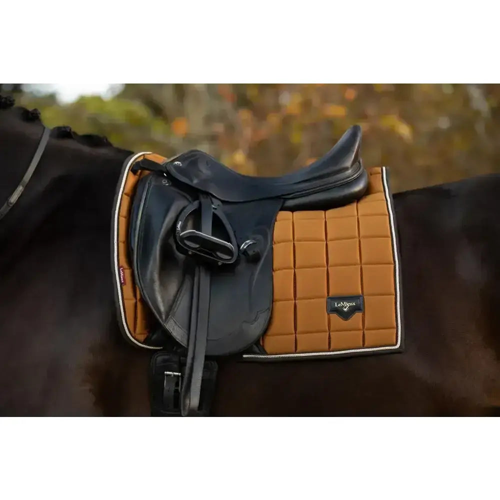Black leather saddle resting on a tan quilted lemieux loire classic dressage square ginger pad with white trim