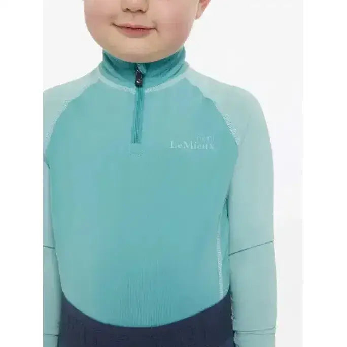 A child wearing a teal lemieux mini base long-sleeved half-zip base layer lagoon with ribbed texture and lamia logo