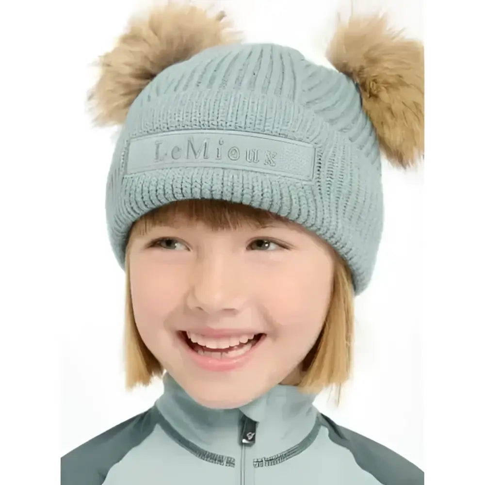 Light blue knitted lemieux mini clara beanie glacier with two tan fur pom-poms and embroidered patch front