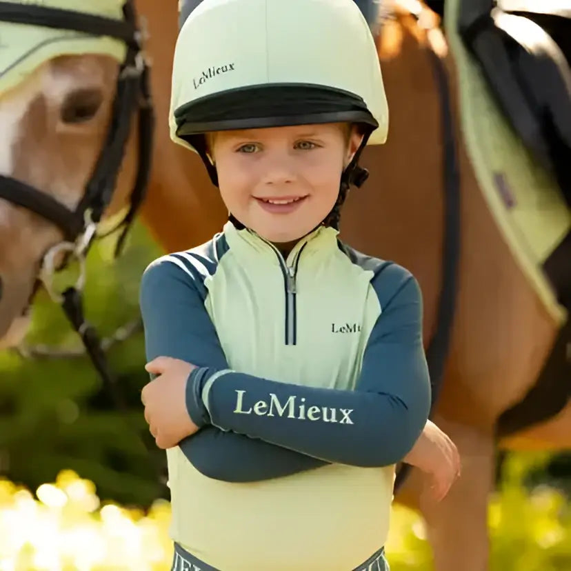 Child wearing a cream lemieux mini silk macaron equestrian helmet with black brim and blue faux fur pom-poms