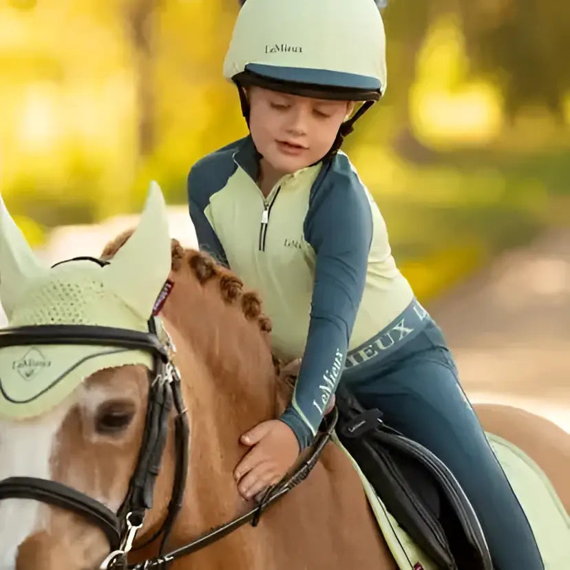 A child wearing a cream lemieux mini silk macaron equestrian helmet with navy blue faux fur pom-poms on top