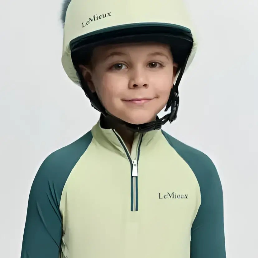 A child wearing a lemieux mini equestrian helmet in pale green and dark teal with black chin strap and faux fur pom silk macaron