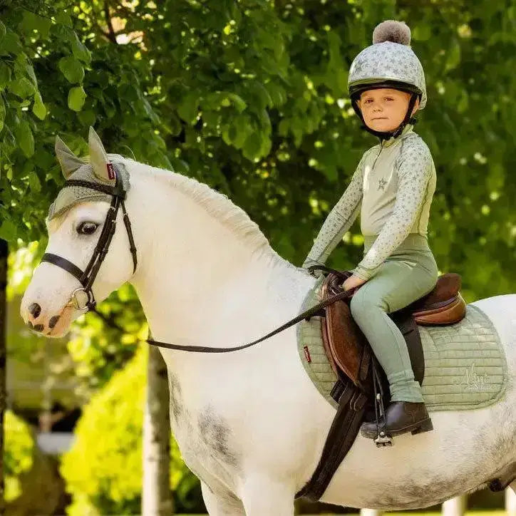 A young rider wearing a lemieux mini fly hood fern in green sits on a white horse with dark markings for riding