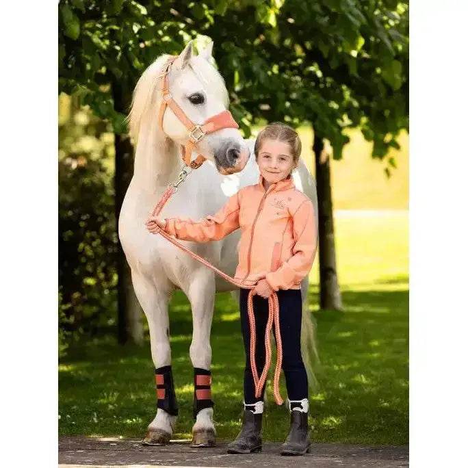 A young girl in a peach jacket stands with a white horse wearing lemieux mini grafter boots apricot with pink lead rope