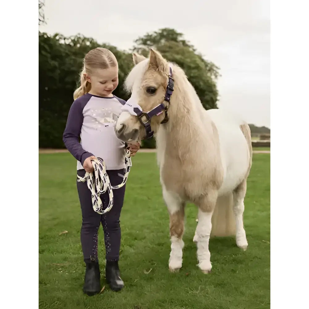A young girl wearing a lemieux mini jamie lemon long sleeve top lilac stands beside a cream pony holding its lead rope