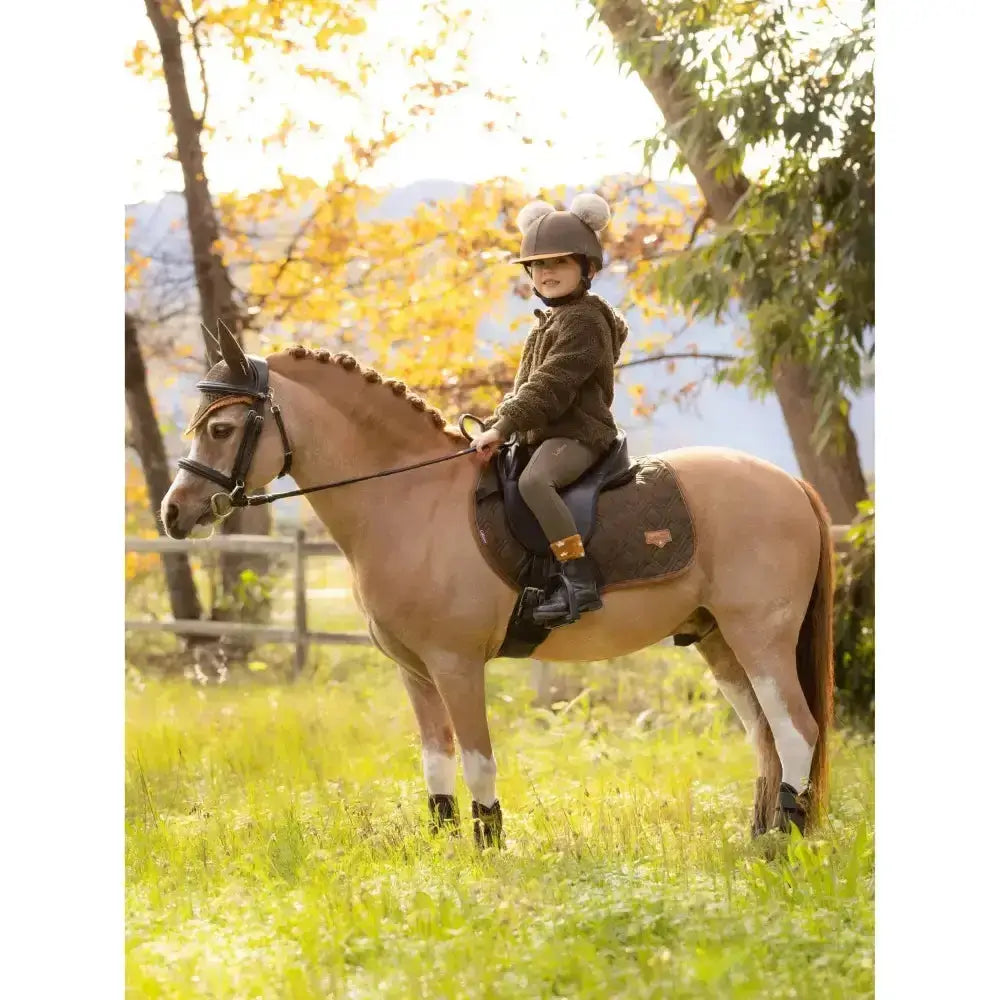A young child wearing a brown helmet rides a light tan horse in an autumn field with lemieux mini pull ons alpine