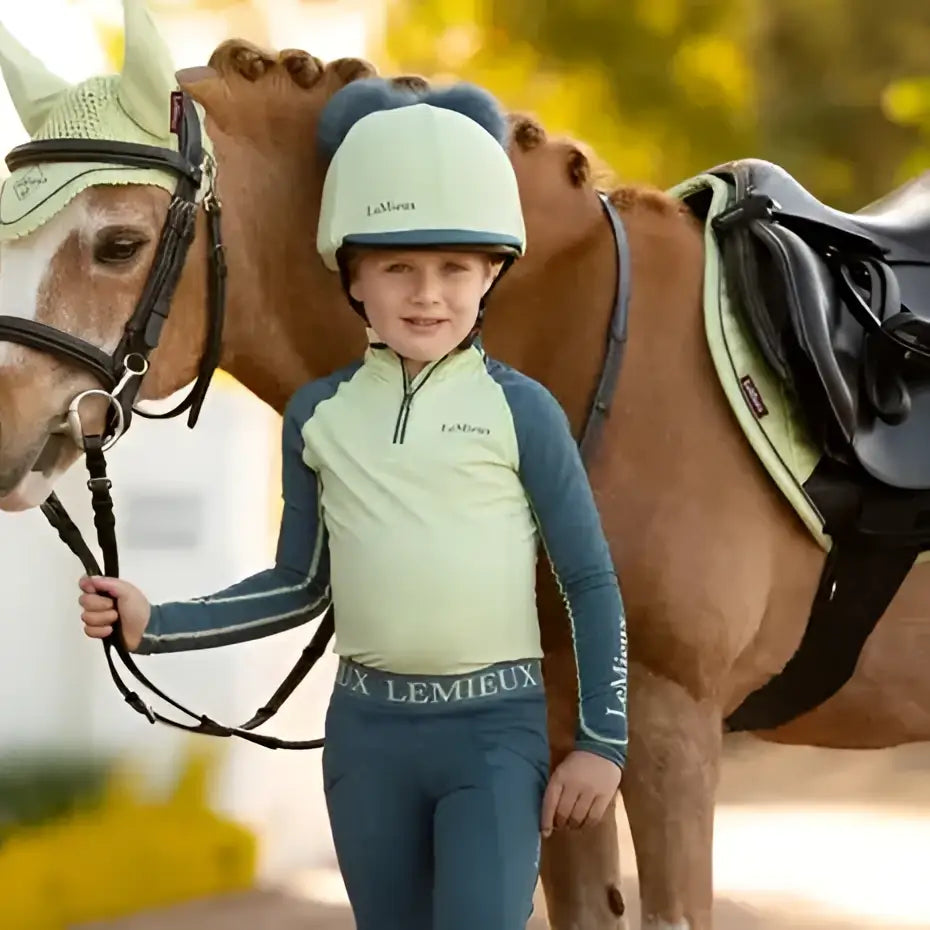 Child wearing lemieux mini suede square macaron with high wither cut in light green and navy l’équipe equestrian outfit and helmet