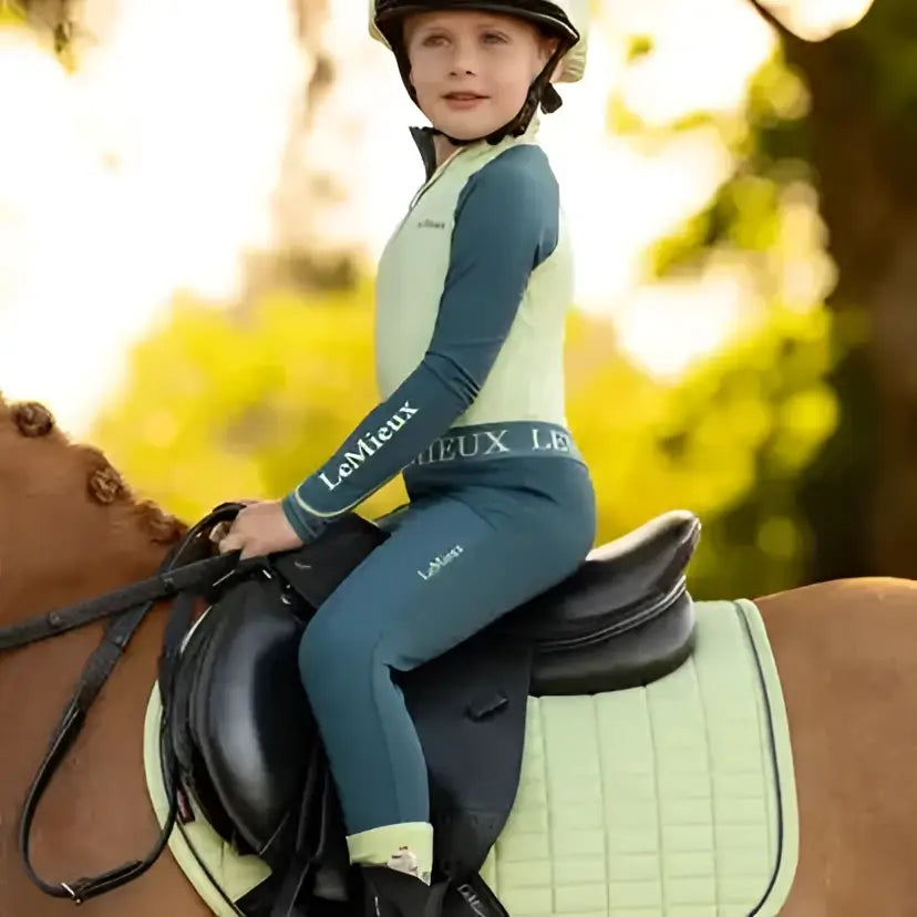 Child wearing a light green and navy blue lemieux mini suede equestrian helmet with high wither cut square macaron design