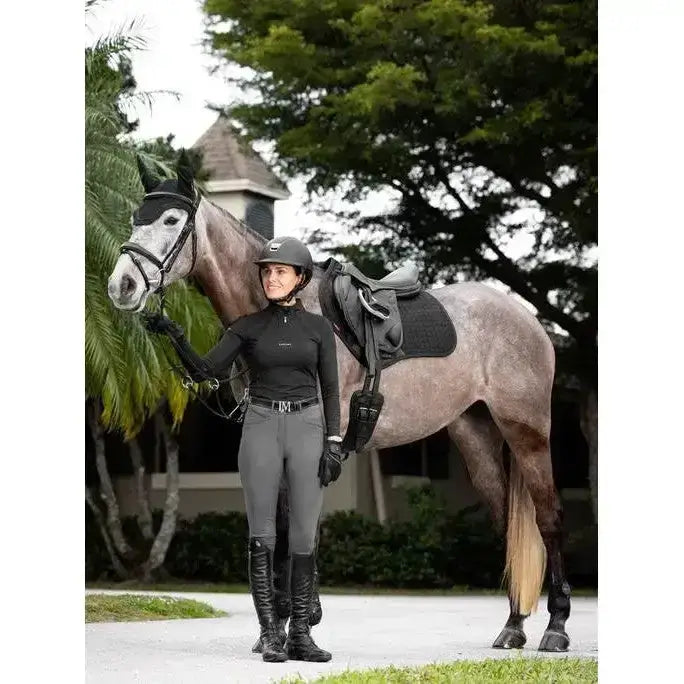 A woman in black riding gear stands beside a gray horse with a contact square black lemieux prosorb plain saddle pad