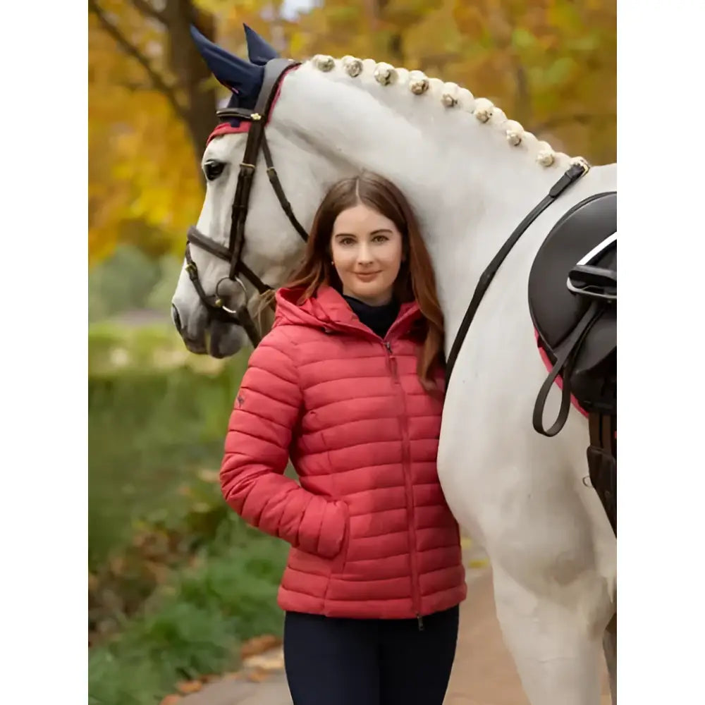 A woman stands beside a white horse wearing a lemieux rose hooded cranberry puffer jacket with front pockets and hood
