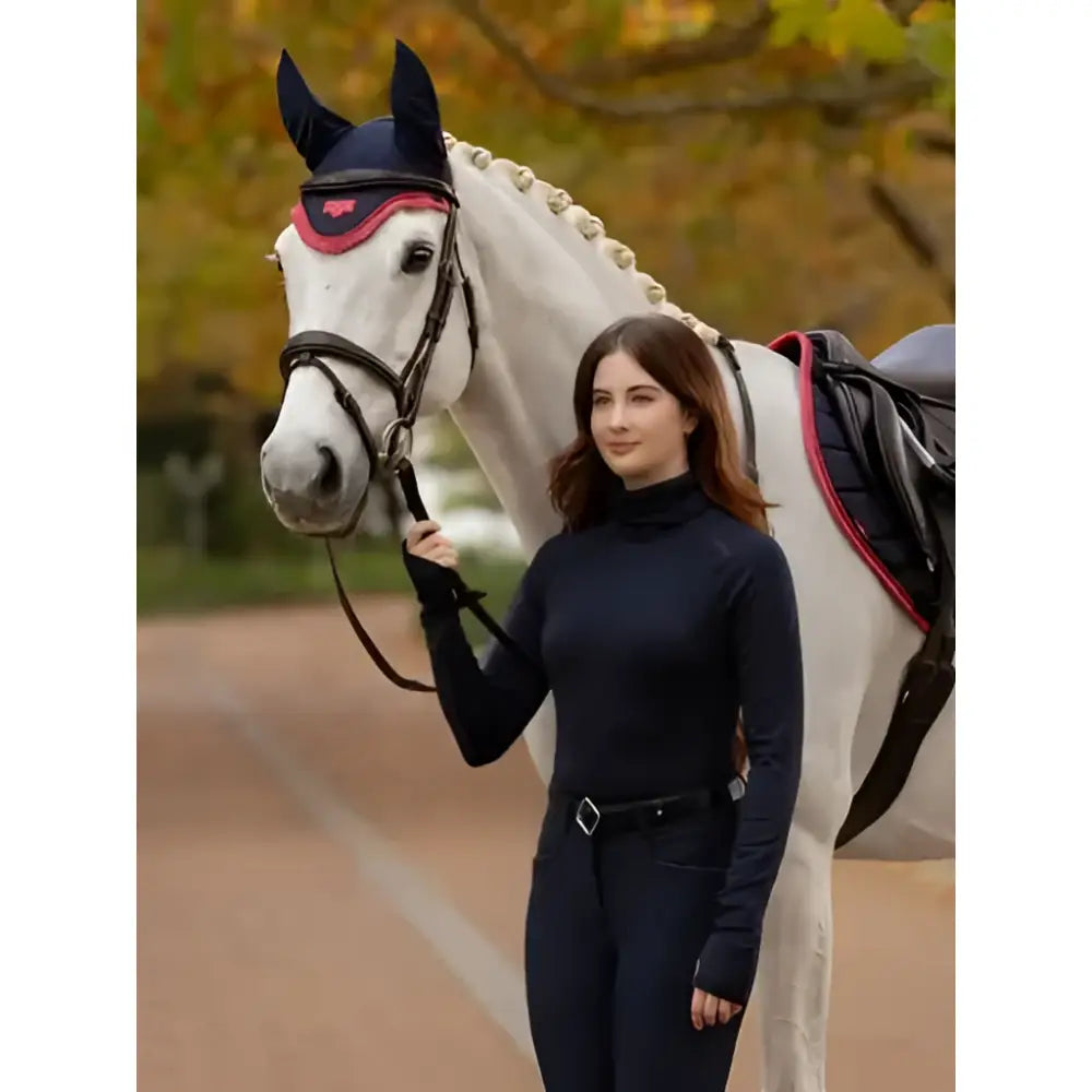 A woman in a navy blue lemieux sara snood base layer stands beside a white horse with a black and red bridle