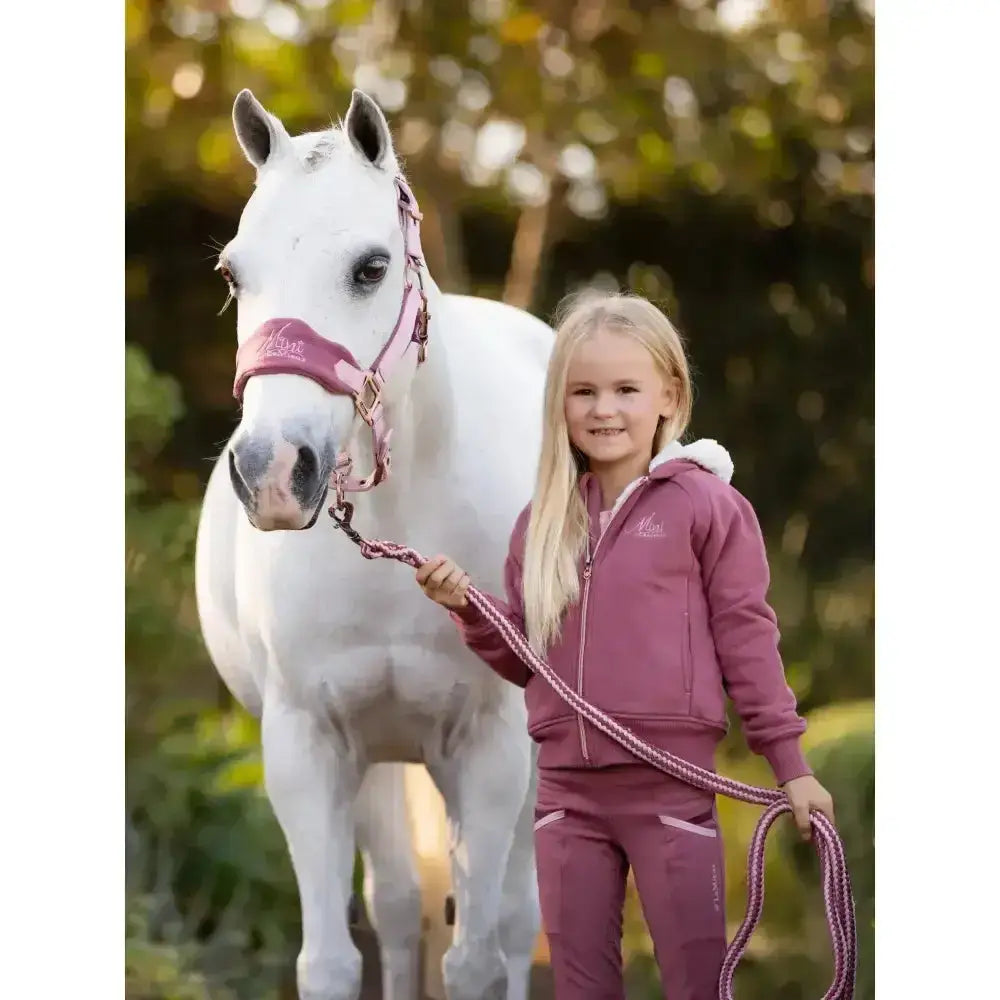 A young girl wearing the lemieux sherpa lined lily hoodie orchid in mauve stands beside a white horse holding its lead rope