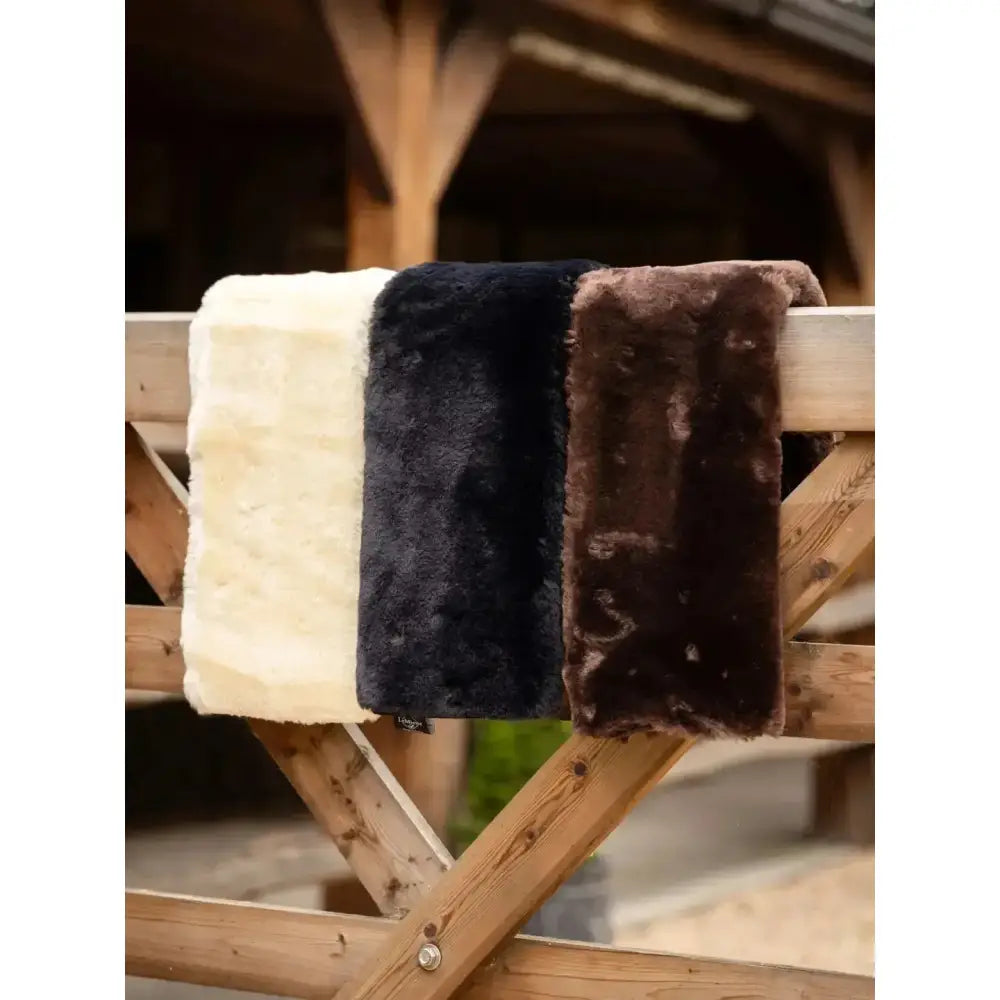 Three cushioned faux lambswool lemieux simuwool dressage girth sleeve blankets in cream black and brown on wooden rack