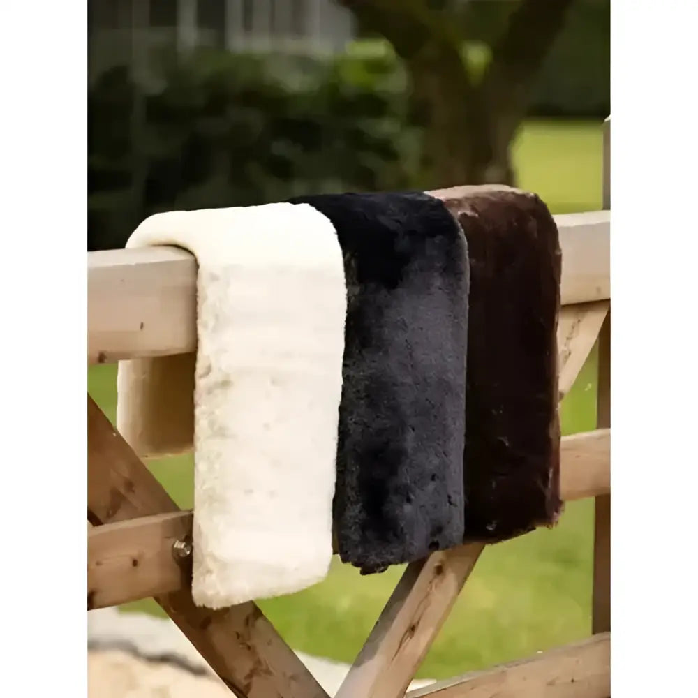 Three lemieux simuwool dressage cushioned faux lambswool girth sleeve natural scarves in cream black and dark brown hanging on wood fence