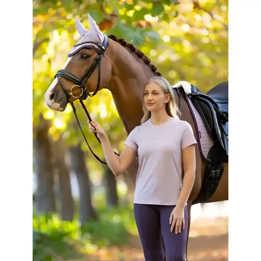 A woman wearing a lemieux sports lilac sports t-shirt stands beside a brown horse with a white mesh eye cover