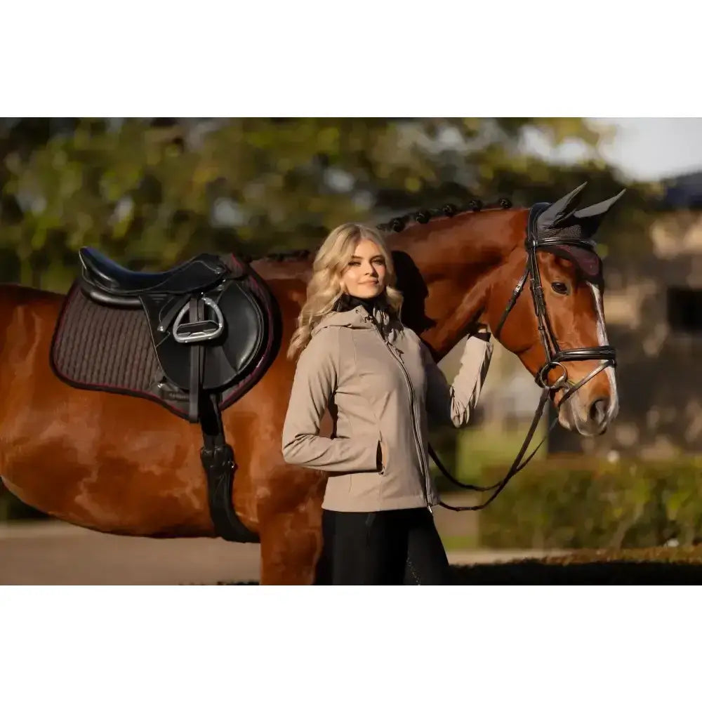 A woman in a beige riding jacket stands beside a brown horse wearing black tack and lemieux suede close contact square cinder saddle pad