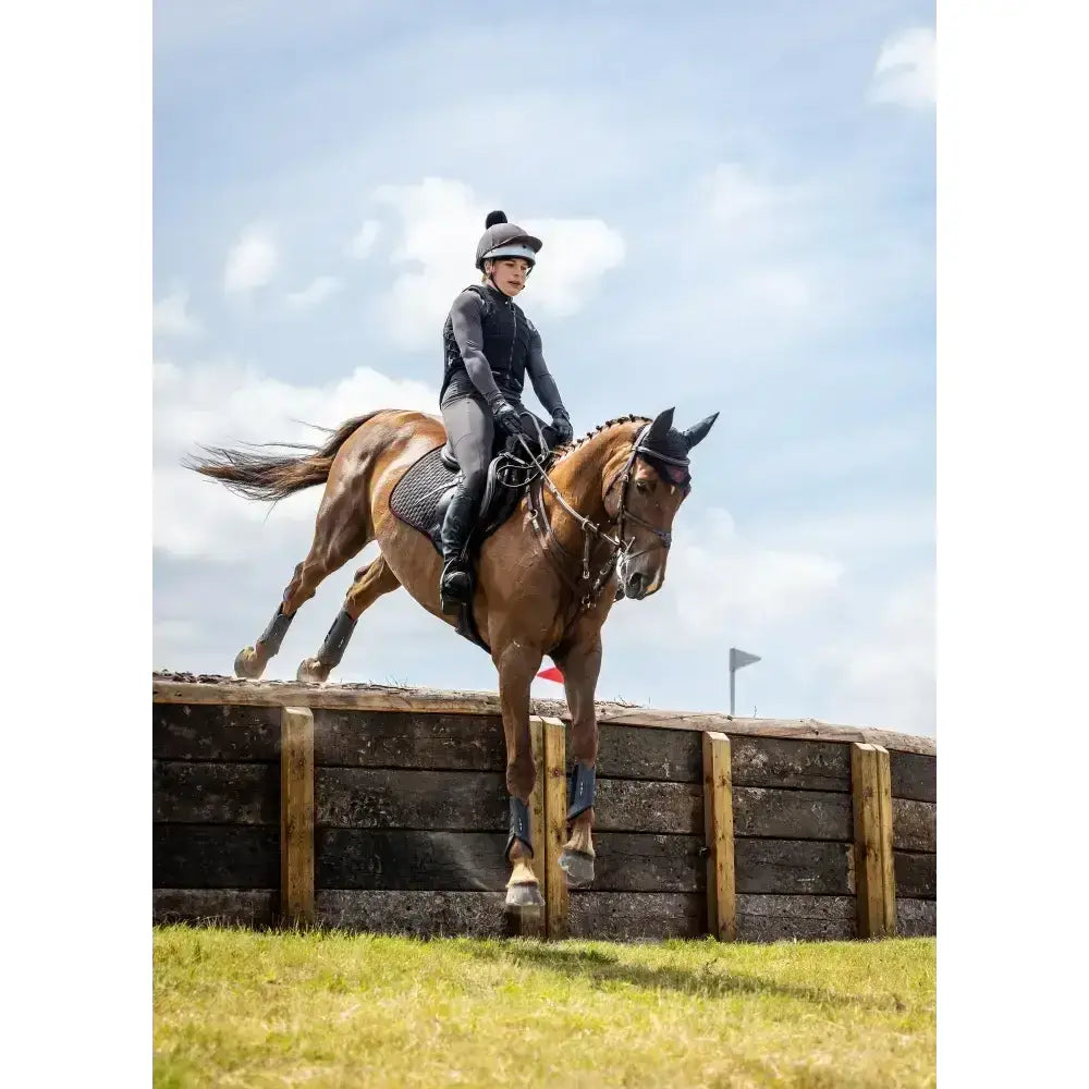 A brown horse with black protective gear and rider mid-jump showcasing lemieux suede close contact square cinder saddle pad