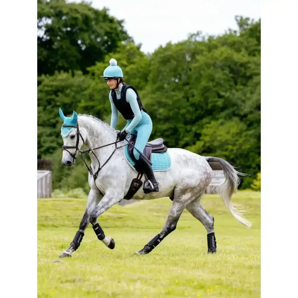 A rider in light blue gear sits on a dapple grey horse wearing matching turquoise lemieux suede close contact square lagoon pads