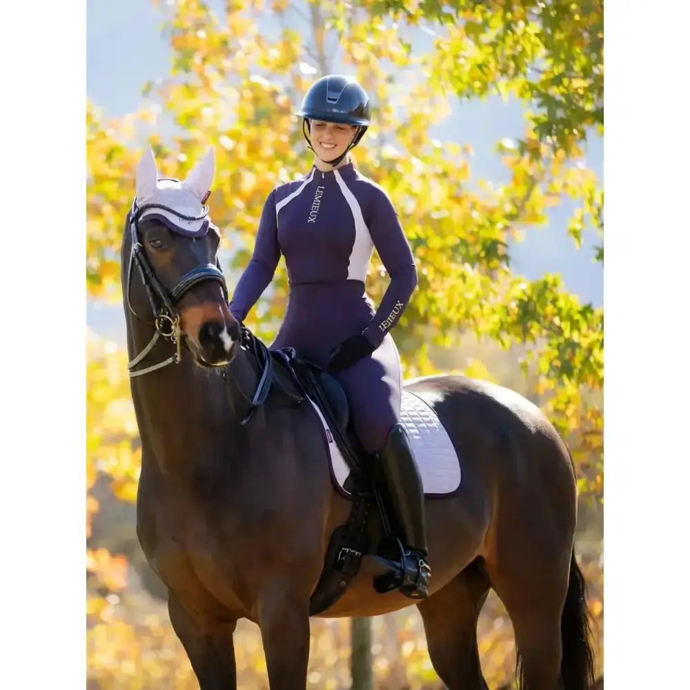 A dark brown horse wearing a lemieux suede dressage lilac saddle pad with a white head cover and black bridle