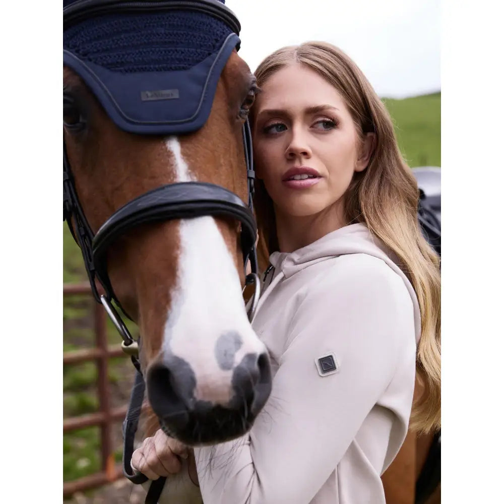 A woman wearing a lemieux tabi tech zip hoodie stone in light beige stands next to a brown horse in navy equestrian gear