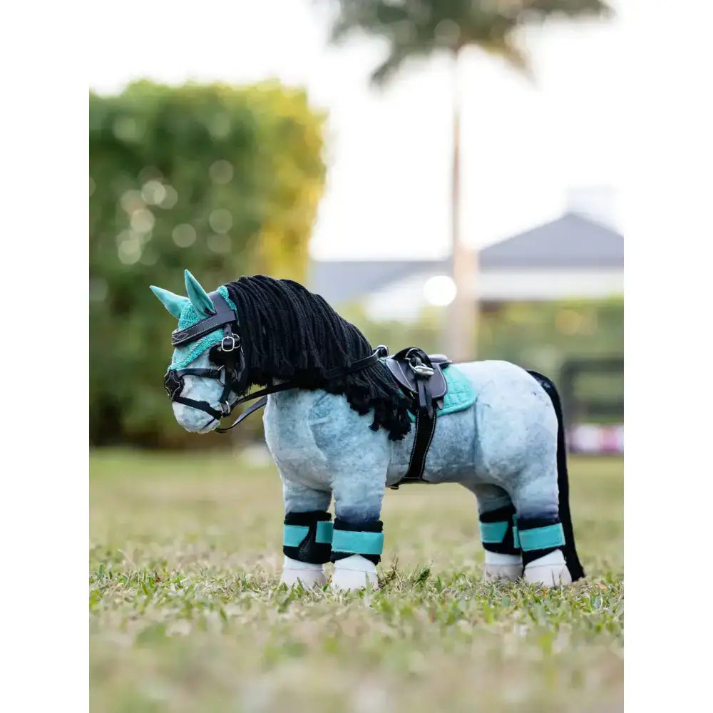 A plush light blue lemieux toy pony with black yarn mane and tail wearing a pony pad lagoon and replica lemieux saddle