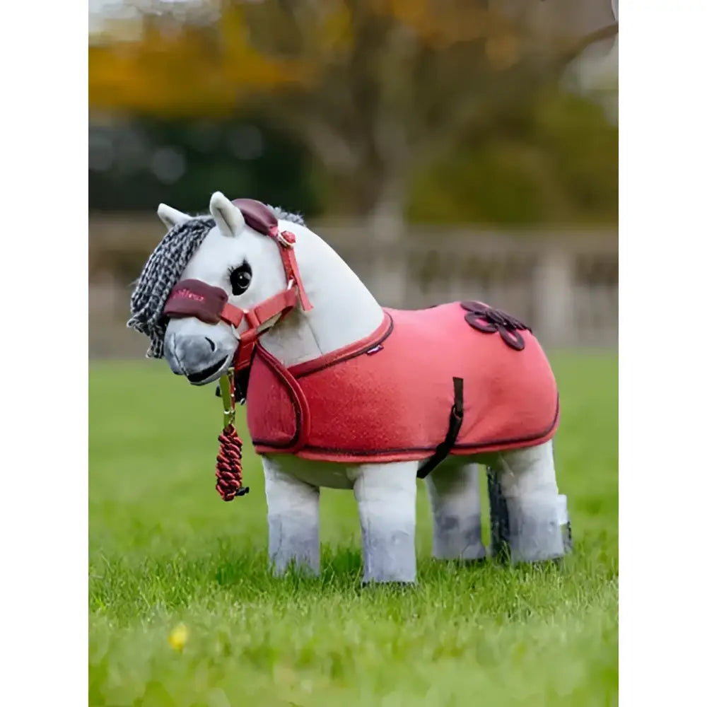 A plush white lemieux toy pony wearing a coral pony rug cranberry fleece rug with black straps and brown bows