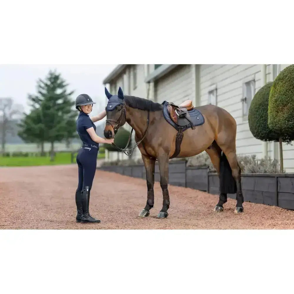 A brown horse stands with a rider wearing equestrian gear using a lemieux ultra mesh close contact square dark navy saddle pad