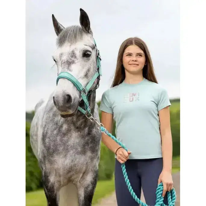 A young girl wearing a lemieux young rider arianna t-shirt aqua stands beside a grey dapple horse with teal halter and lead rope